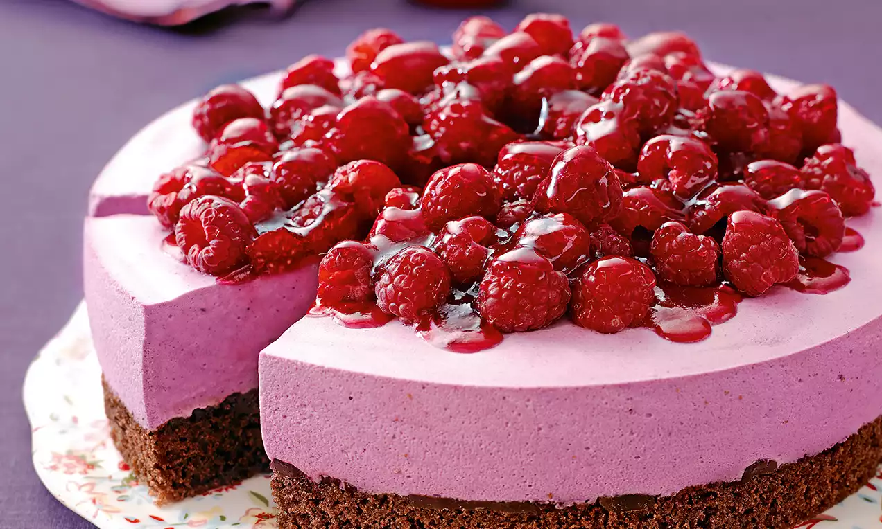 Beeren-Mousse-Torte mit satter, violetter Creme auf dunklem Schokoboden, bedeckt von glänzenden Himbeeren auf weißem Porzellanteller