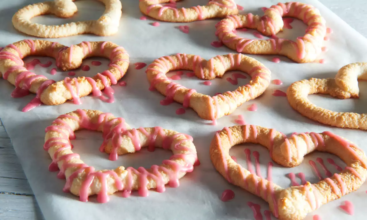 Svijetložuti kolačići u obliku srca s hrskavom teksturom, ukrašeni nepravilnim ružičastim preljevom, posloženi na bijelom papiru za pečenje