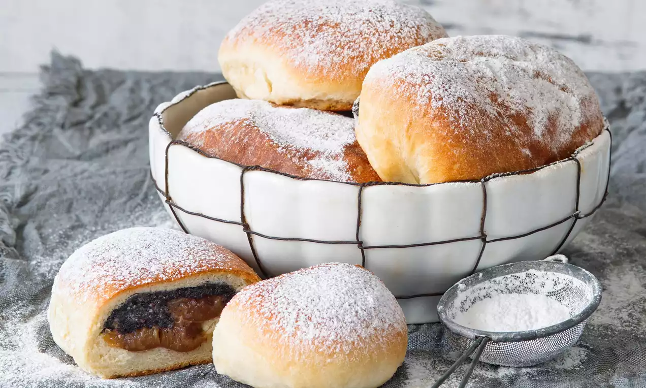 Nadýchané kulaté pečivo zlatohnědé barvy, uvnitř tmavá maková náplň, uspořádané v bílé míse, posypané jemným moučkovým cukrem