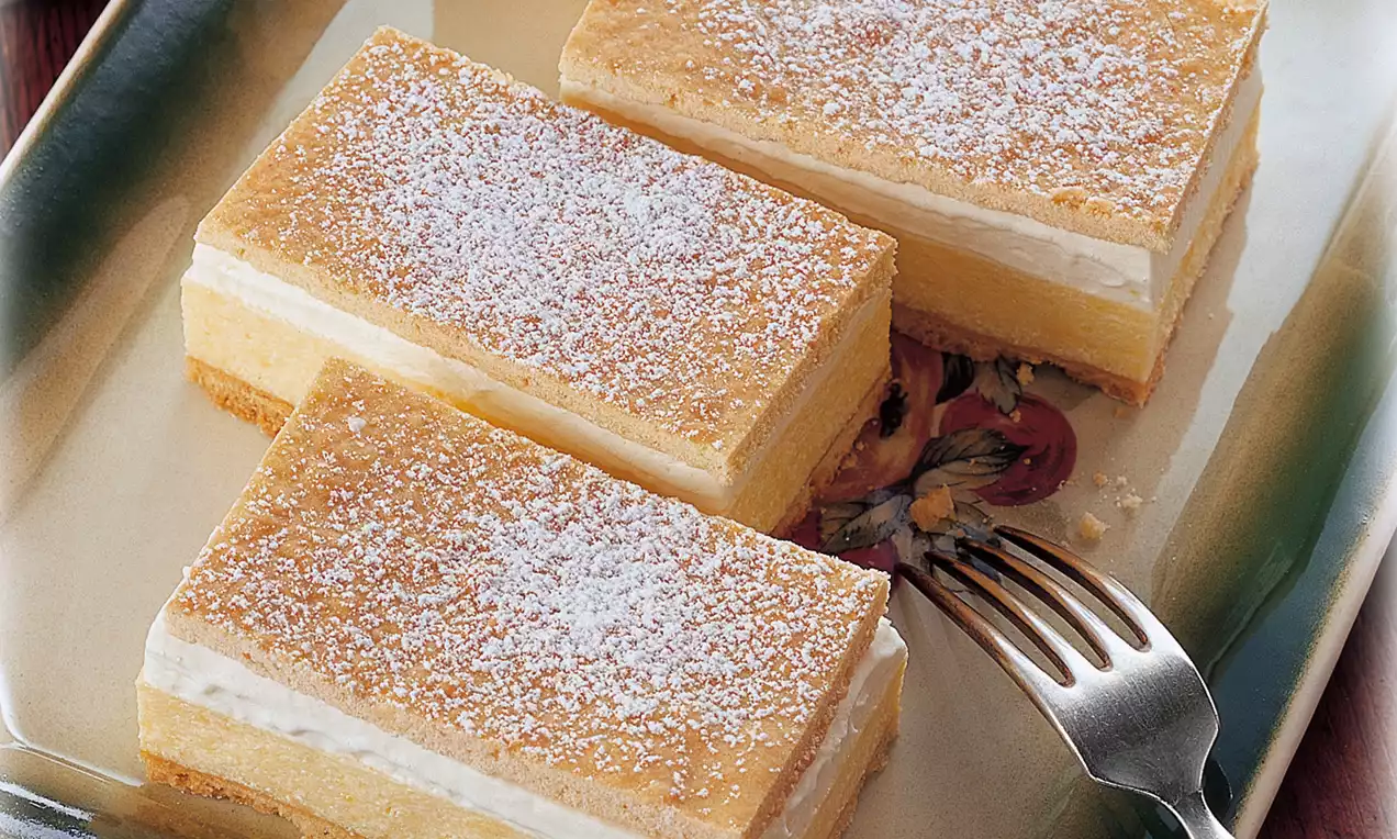 Rechteckige Cremeschnitten mit heller Schichtfüllung, goldgelber Teigdecke und Puderzucker bestäubt auf einem Teller mit Gabel