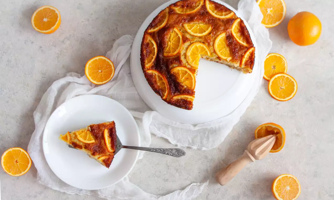 Portokalopita aurie, cu textură umedă și felii de portocală caramelizate dispuse simetric, pe un platou alb și o farfurie cu șervețel de pânză albă