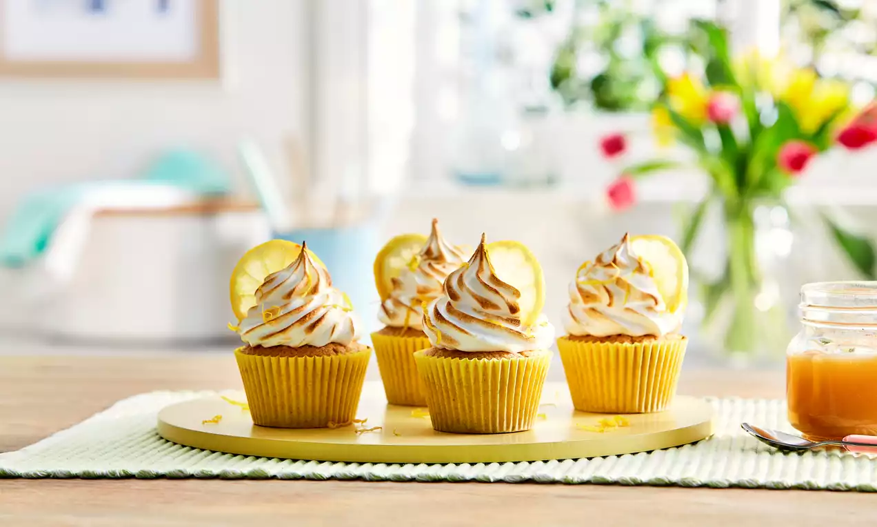Warm golden cupcakes with swirled toasted meringue peaks, each topped with a slice of lemon, arranged on a pale yellow plate on a table