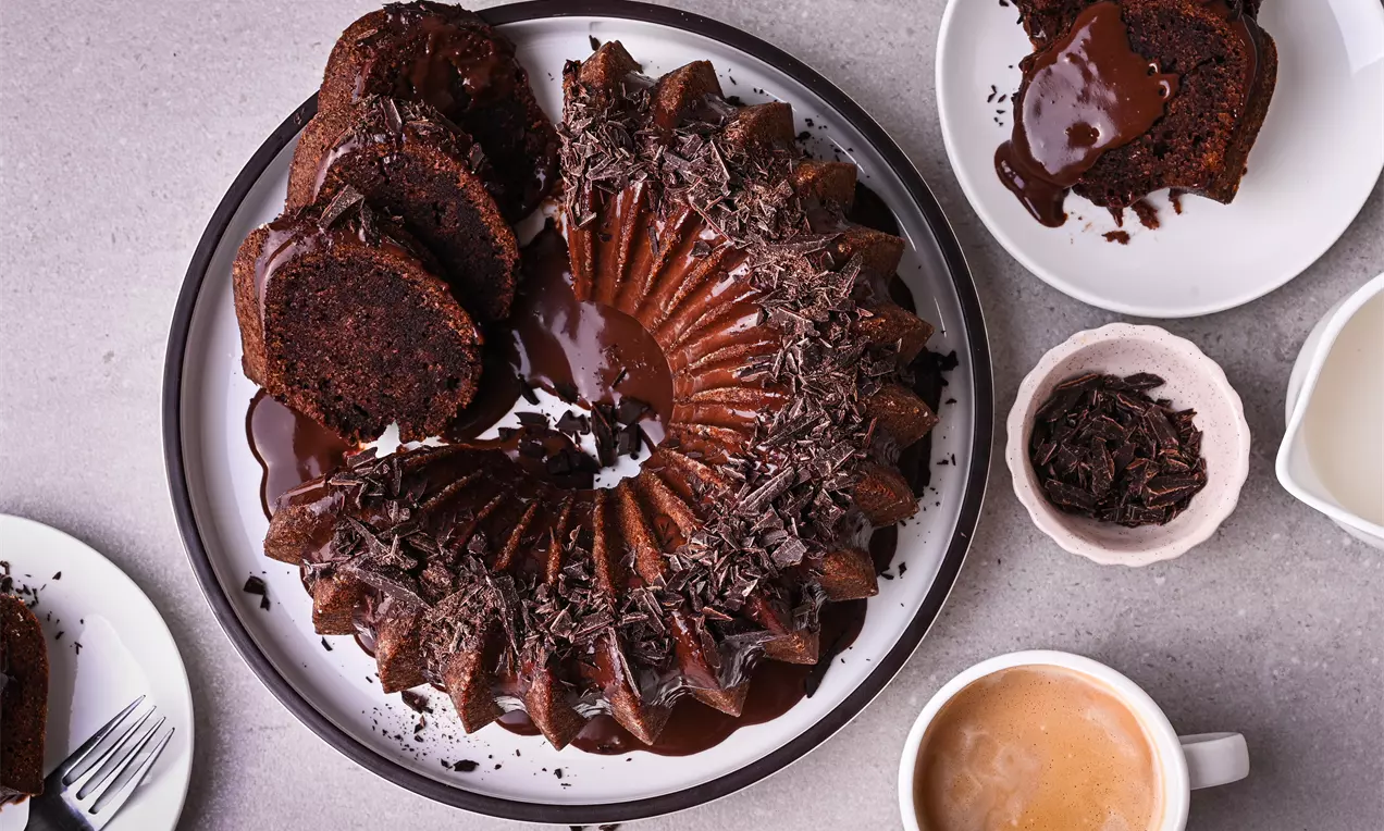 Шоколадов кекс във форма на венец с гланцова шоколадова глазура, декориран с настърган шоколад, сервиран на бяла чиния