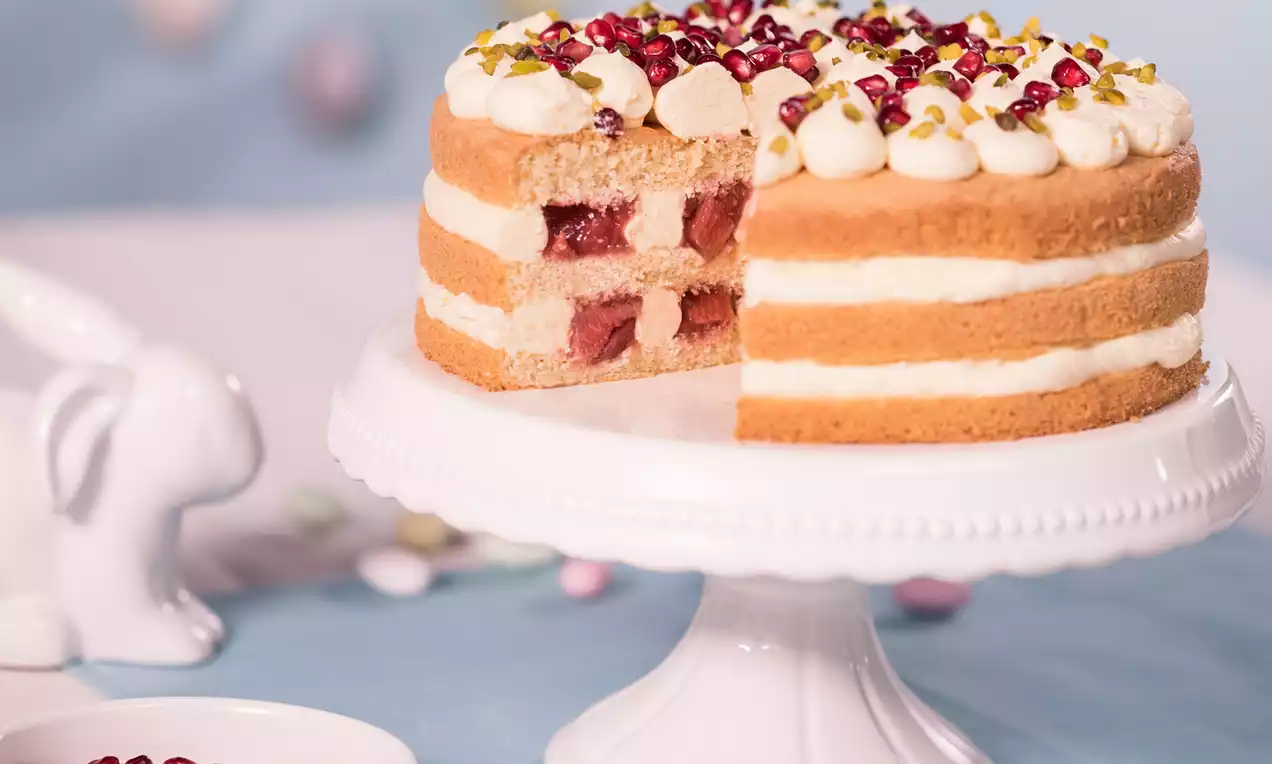 Naked cake met lagen goudbruine biscuit, witte room, rabarbervulling en decoratie van granaatappelpitjes en pistachenoten op een wit taartplateau