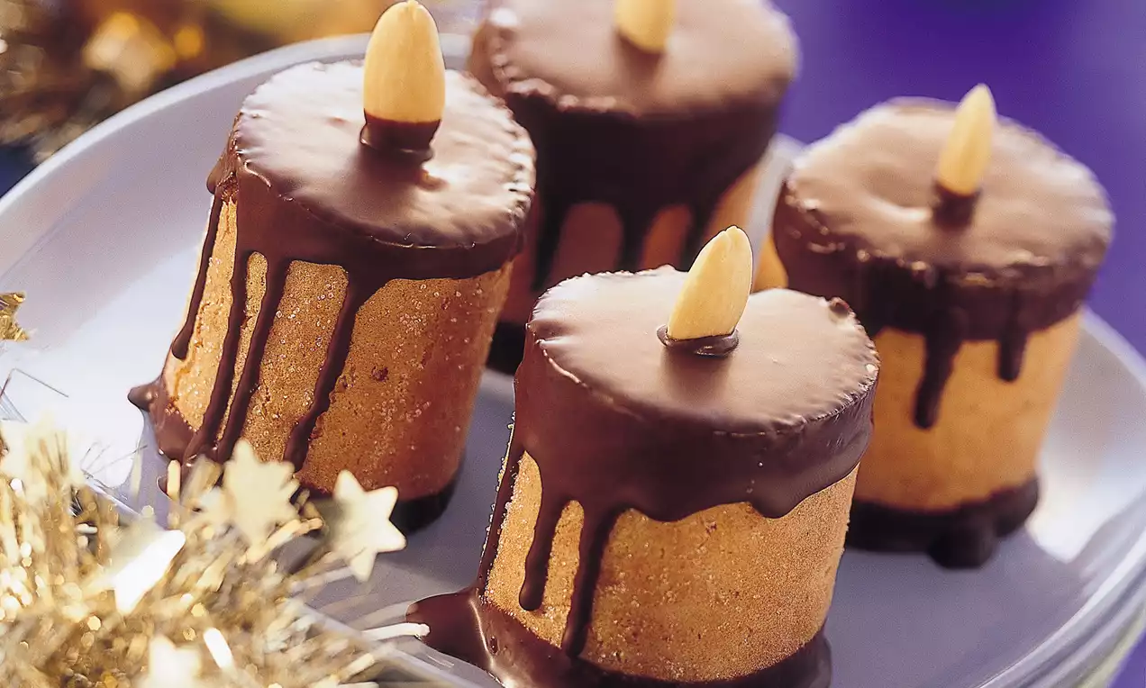 Vier zylinderförmige Süßgebäcke mit schokoladenbrauner Glasur und Mandeldekor, aufgestapelt auf lilafarbenem Teller, umgeben von goldenen Kugeln