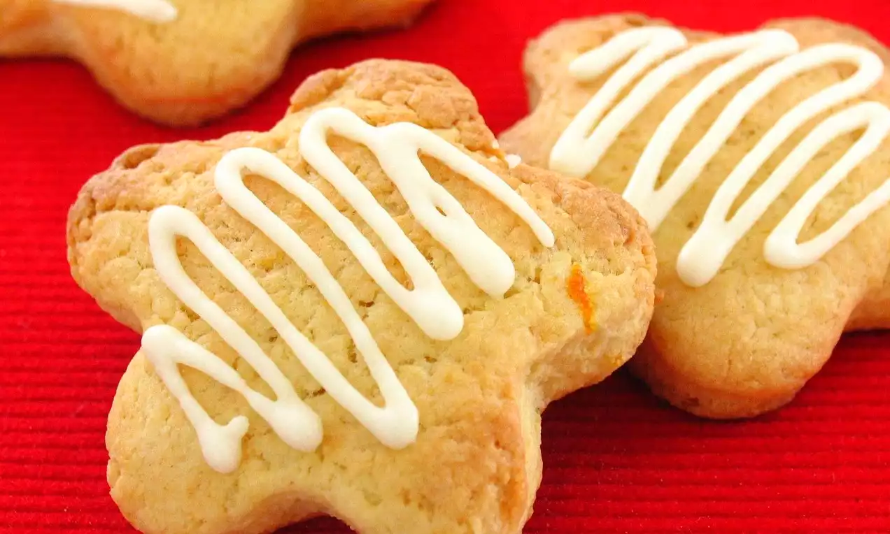 Portakal aromalı kurabiyeler altın renginde, yıldız şeklinde, üzerlerinde beyaz şekerli glazür desenleri ile kırmızı zemin üzerinde sergilenmiş