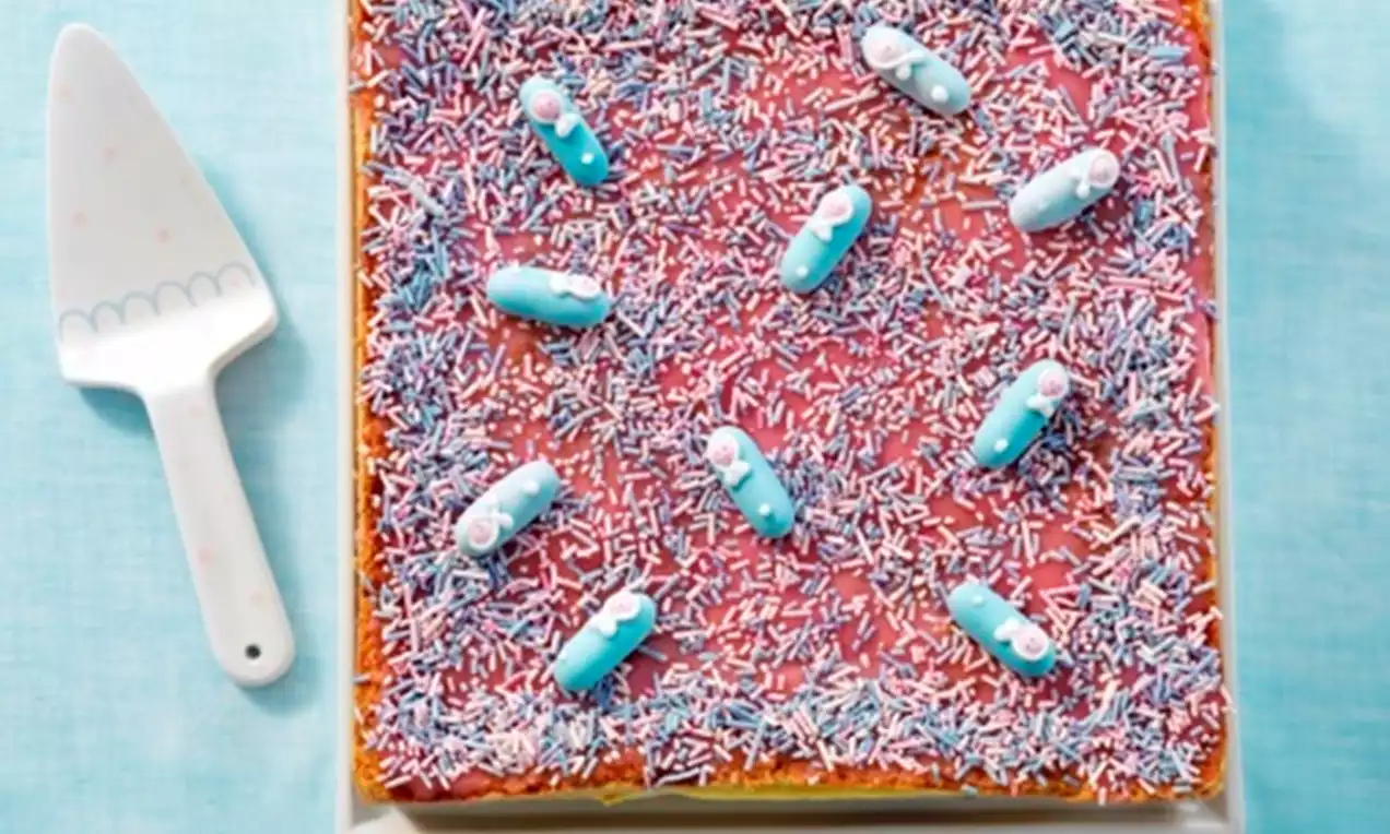 Vierkante geboortetaart met roze glazuur, blauwe snoepjes en wit-blauwe muisjes op een wit bord met een taartschep ernaast