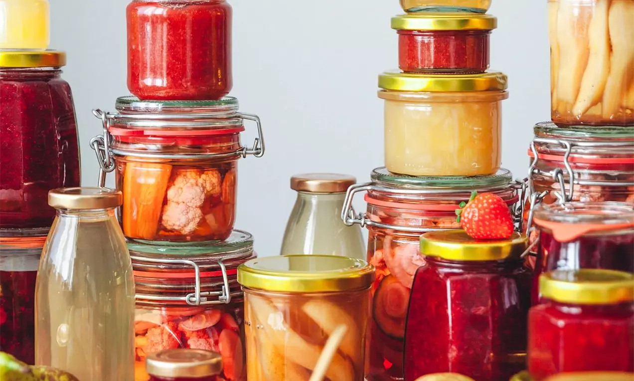 Pestrofarebné zavařeniny v sklenených pohároch rôznych tvarov s kovovými viečkami, usporiadané na drevenom stole, obklopené čerstvým ovocím