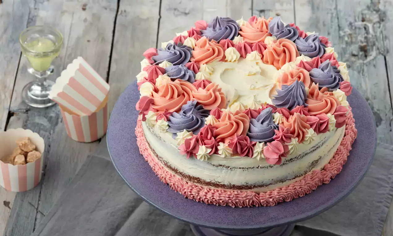 Runde Torte mit weißer Sahneschicht und rosa, lila und cremefarbenen Blütentupfern auf violetter Platte, dekoriert mit Bechern und Glas