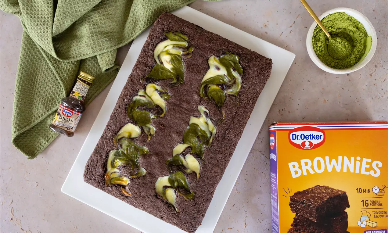 Donkergroene matcha-swirls in een rechthoekige, donkerbruine brownie op een wit bord met een potje matchapoeder en een groene doek ernaast