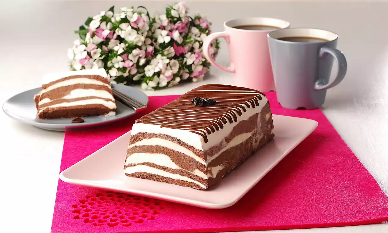 Kakaolu ve beyaz katmanlı dikdörtgen parfe, çikolata şeritleriyle süslenmiş, pembe örtü üzerinde beyaz tabakta sunulmuş