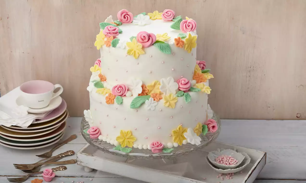 Weiße zweistöckige Hochzeitstorte, verziert mit rosa Rosen, gelben und weißen Blüten sowie grünen Blättern auf Glasplatte und Holzuntergrund