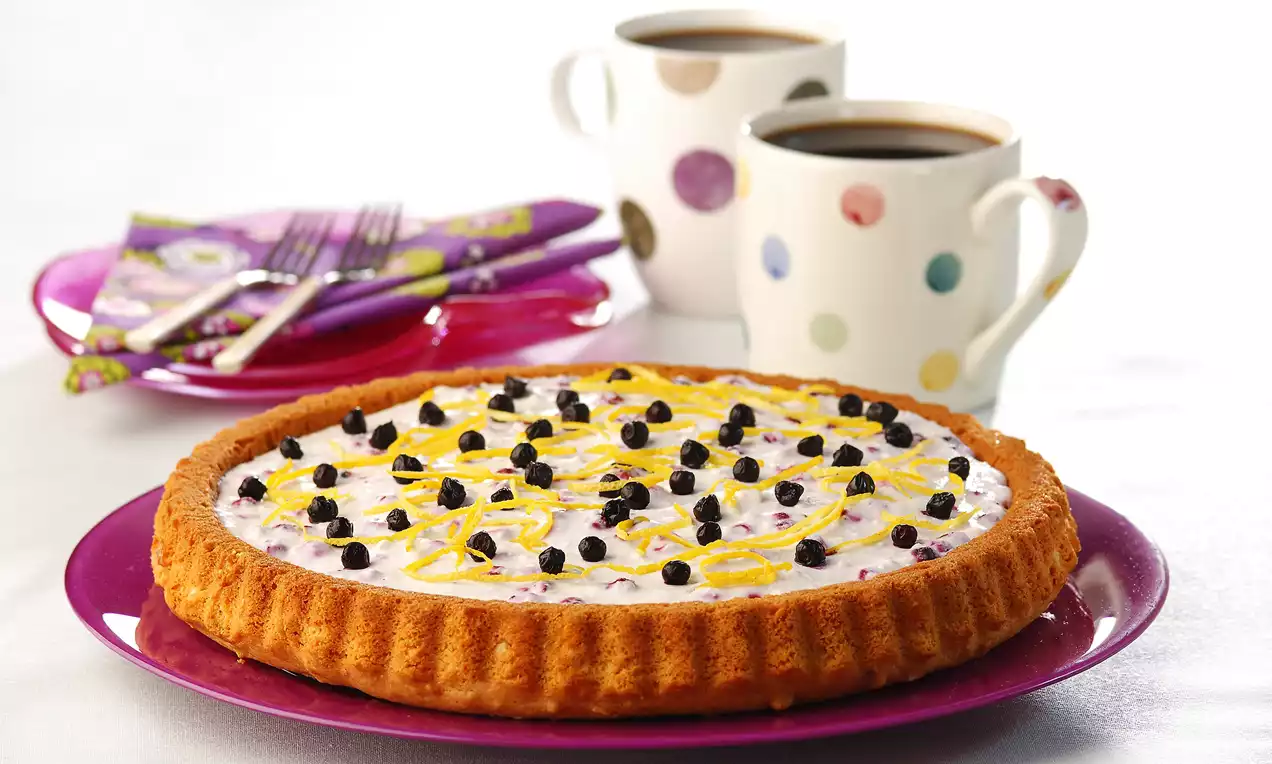 Altın kahverengi tart tabanı üzerinde kremsi dolgu, yaban mersini taneleri ve rendelenmiş limon kabuğu ile mor bir tabakta sunulmuş