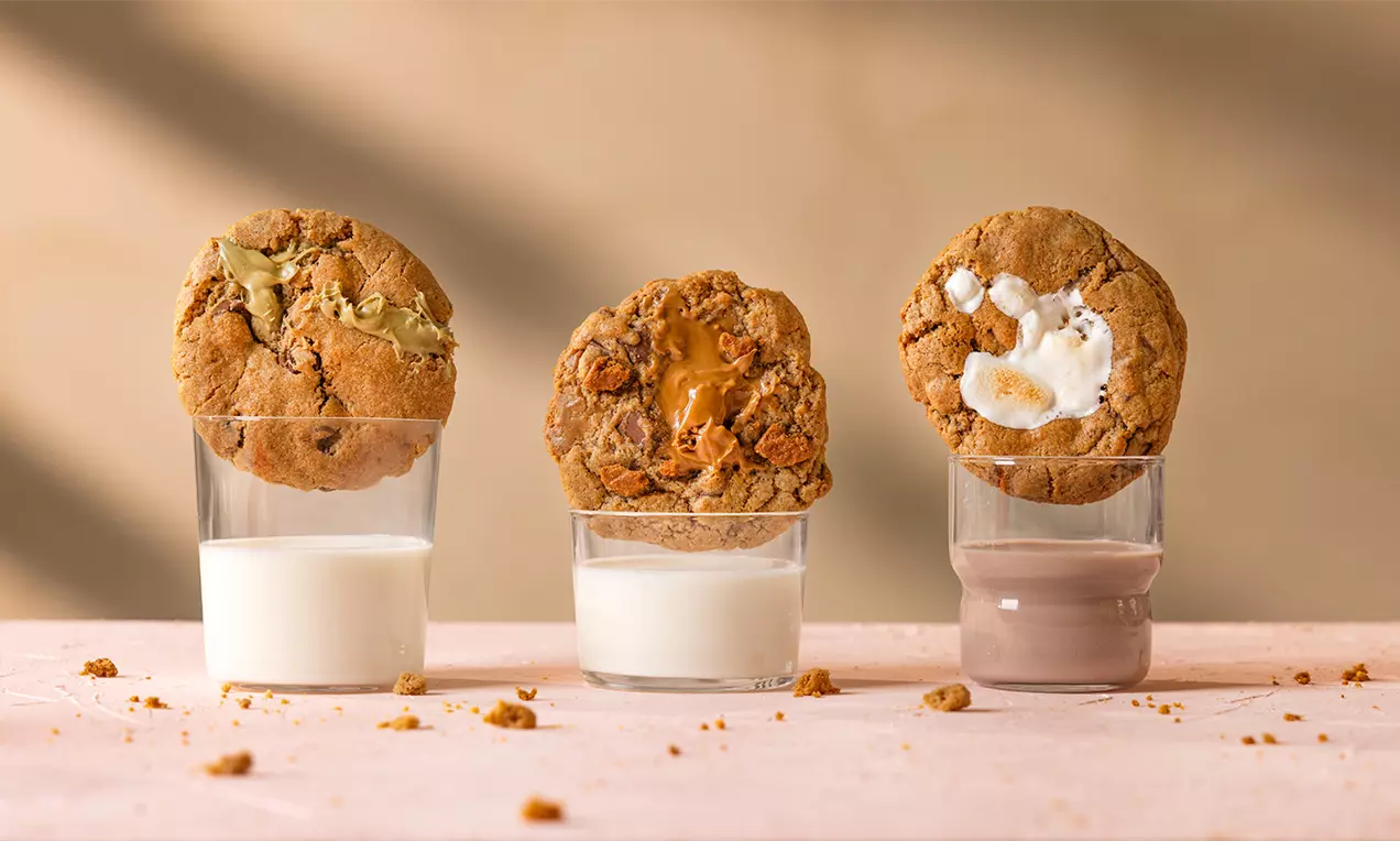 Drie ronde, goudbruine chocolate chip cookies met zachte vulling, rustend op glazen melk, in een warme setting met lichte kruimels