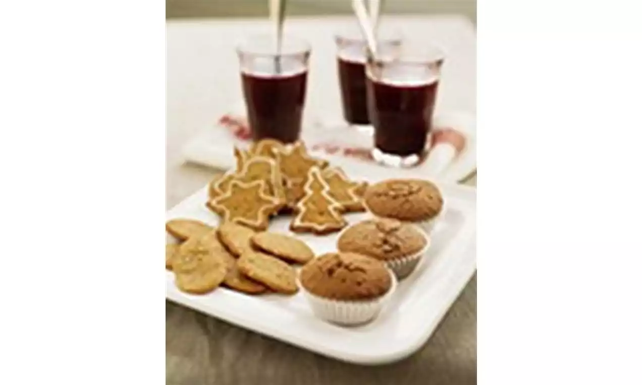 Pepparkakor i olika former och gyllenbruna muffins i vita formar på vit fyrkantig tallrik, med glas röd glögg i bakgrunden