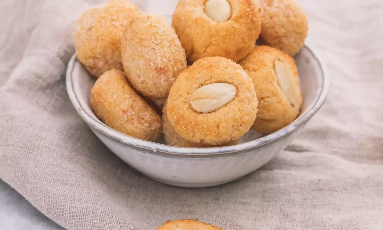 Panellets de almendra dorados, redondeados y de textura granulada, con una almendra entera al centro, servidos en un cuenco cerámico sobre un paño beige
