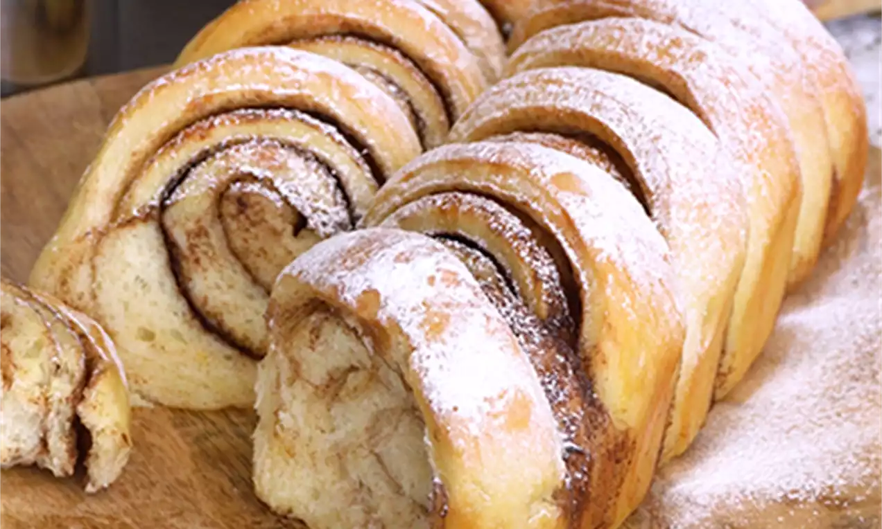 Pahuljaste cimet rolice zlatnosmeđe boje s uvijenom teksturom, poslužene na tanjuru uz štapiće cimeta i bijeli keramički vrč s točkama