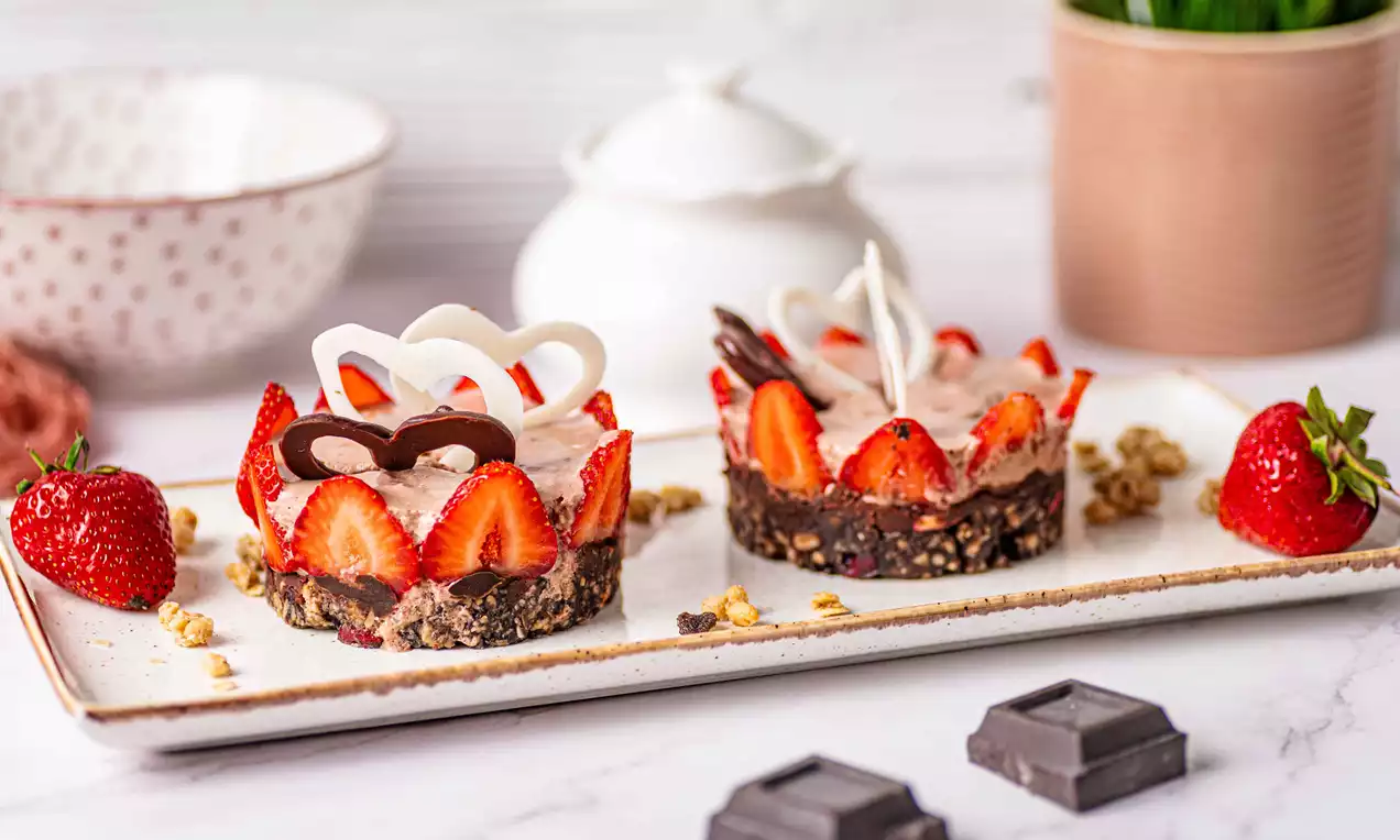 Mini tortulețe rotunde cu blat crocant de musli, decorate cu felii de căpșuni, ornamente de ciocolată și presărate cu granola pe un platou dreptunghiular crem