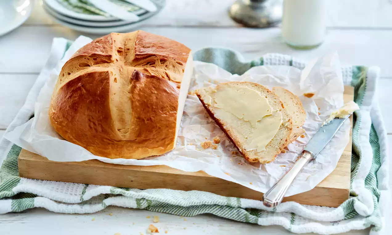 Golden brown round loaf with a cross-shaped cut, resting on parchment paper, beside a slice spread with creamy butter on a wooden board