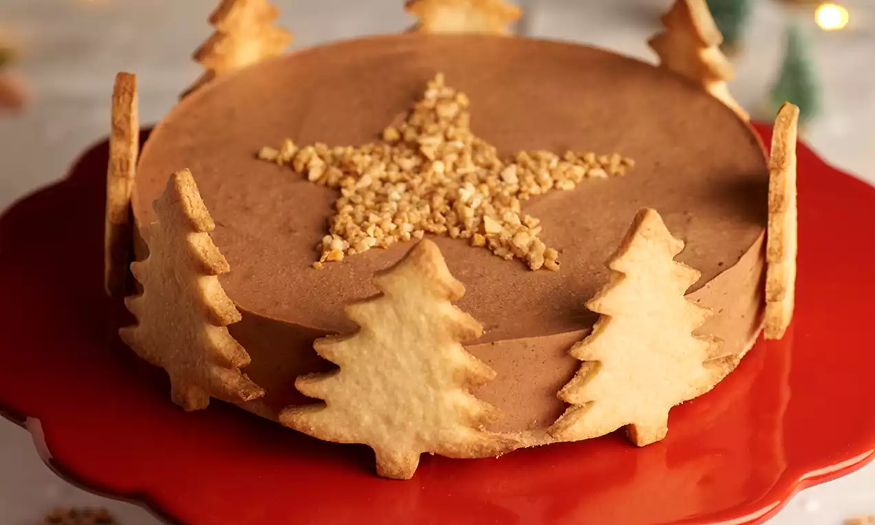 Tarta circular de crema marrón suave decorada con una estrella de crocante en el centro y galletas en forma de abeto alrededor sobre un plato rojo