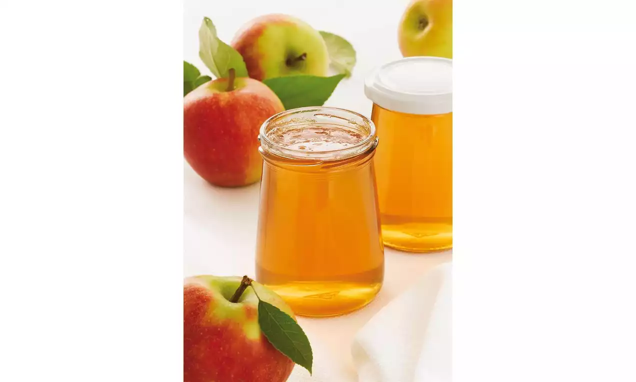 Helder gouden appelgelei in glazen potten met witte deksel, omringd door rode appels met groene blaadjes op een lichte achtergrond