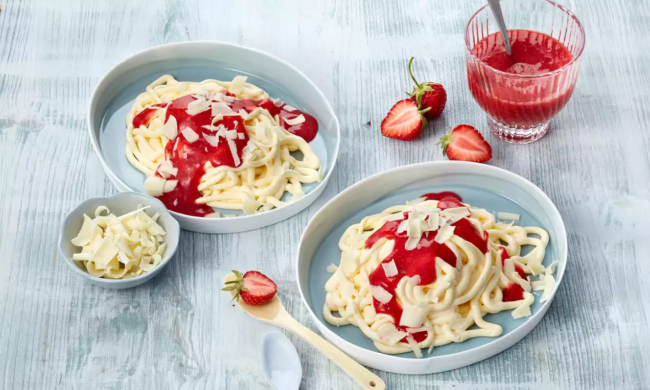Spaghetti-Eis mit weißer Vanillecreme, roter Erdbeersoße und weißen Schokospänen in zwei runden Schalen auf heller Holzoptik