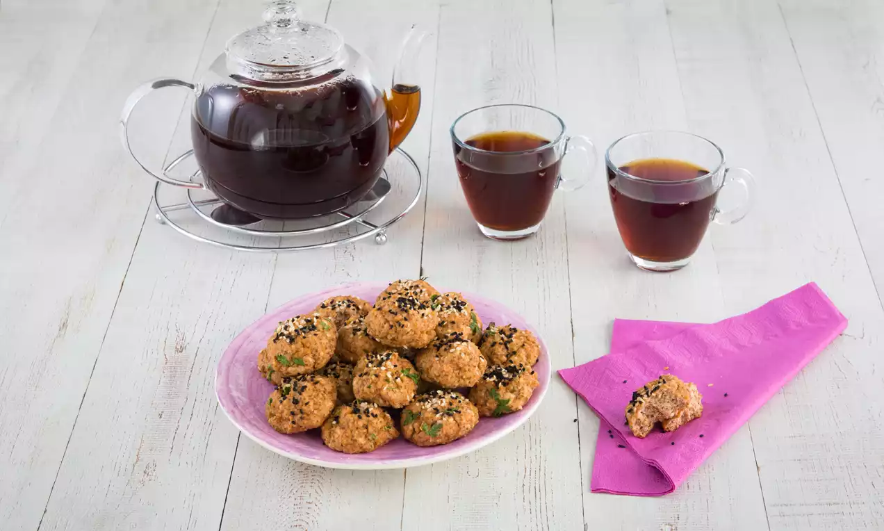 Pembe tabakta sunulan altın rengi unsuz lor peynirli poğaçalar, üzerlerine serpilmiş susam ve çörekotu ile çay eşliğinde düzenlenmiş