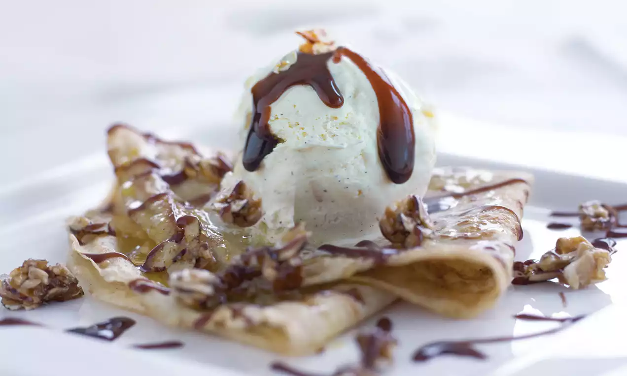 Tynne crêpes brettet i trekanter, toppet med en kule hvit vaniljeis, sjokoladesaus og karamelliserte nøtter på en lys tallerken