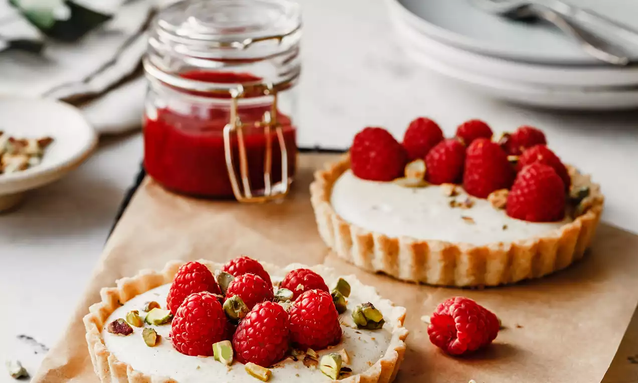 Picture - Golden vanilla tarts with creamy white filling topped with vibrant red raspberries and crushed green pistachios on parchment paper