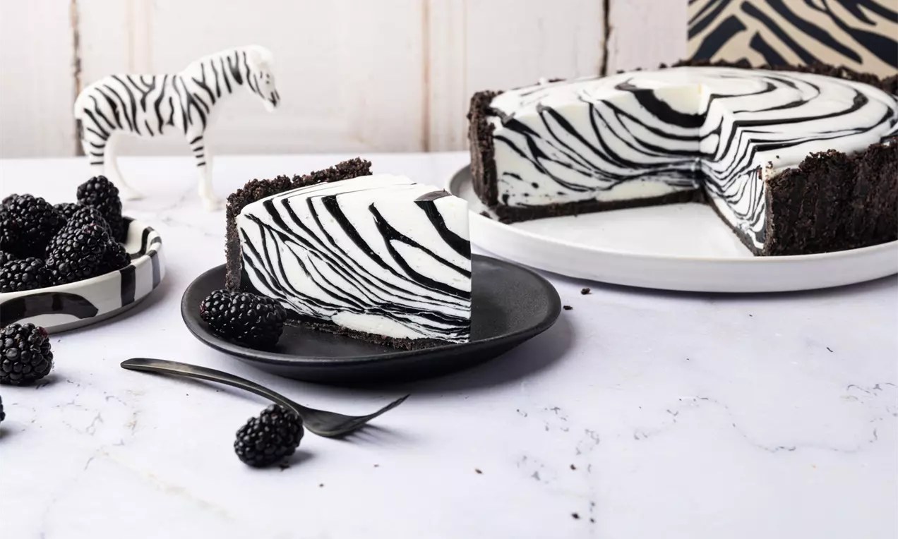 Sernik na zimno w paski czarno-białe z kremowym środkiem i spodem z ciemnych herbatników, podany na białych talerzach ze złotym widelcem