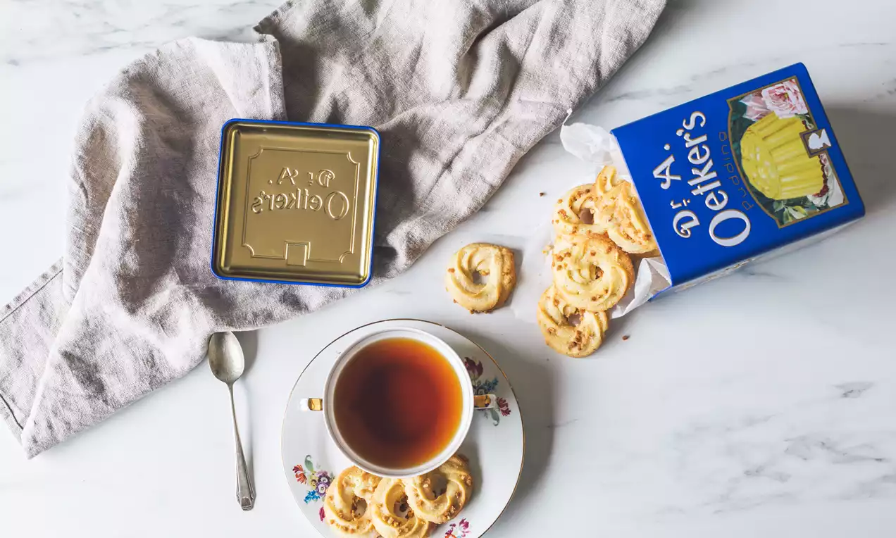 Galletas doradas en espiral con textura crujiente dispuestas en un plato floral junto a una taza de té, una lata y un paño beige