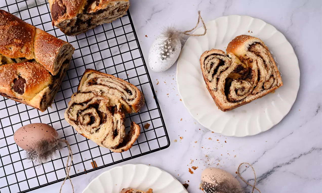 Козунак златисто-кафяв с разлято тесто, вътре слоеве течен шоколад, нарязан на филии върху бяла чиния и решетка