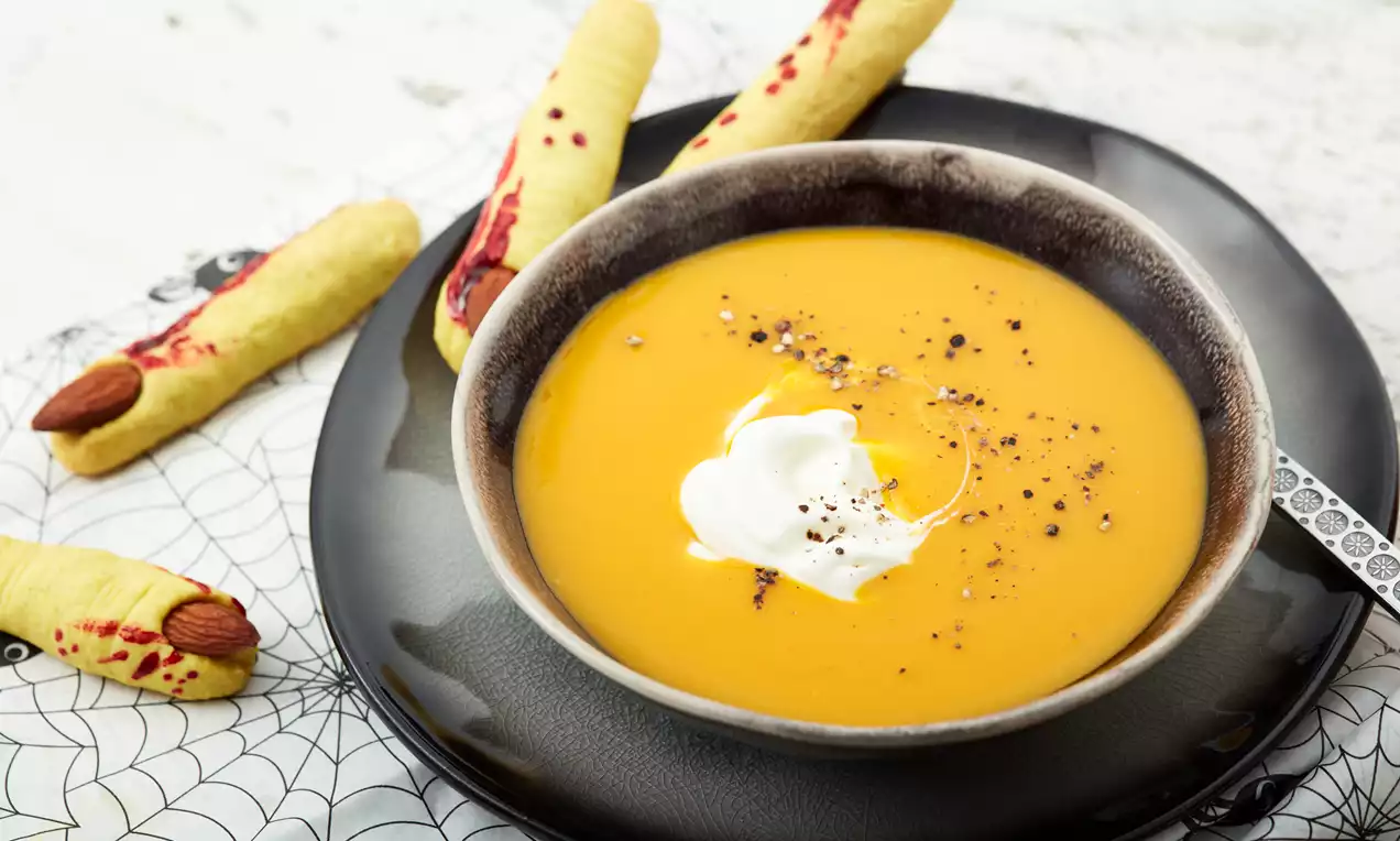 Kürbissuppe in einem dunklen, rundlichen Teller mit Sahnehäubchen und Pfeffer, arrangiert mit gruseligen Hexenfinger-Keksen auf Netz-Deckchen