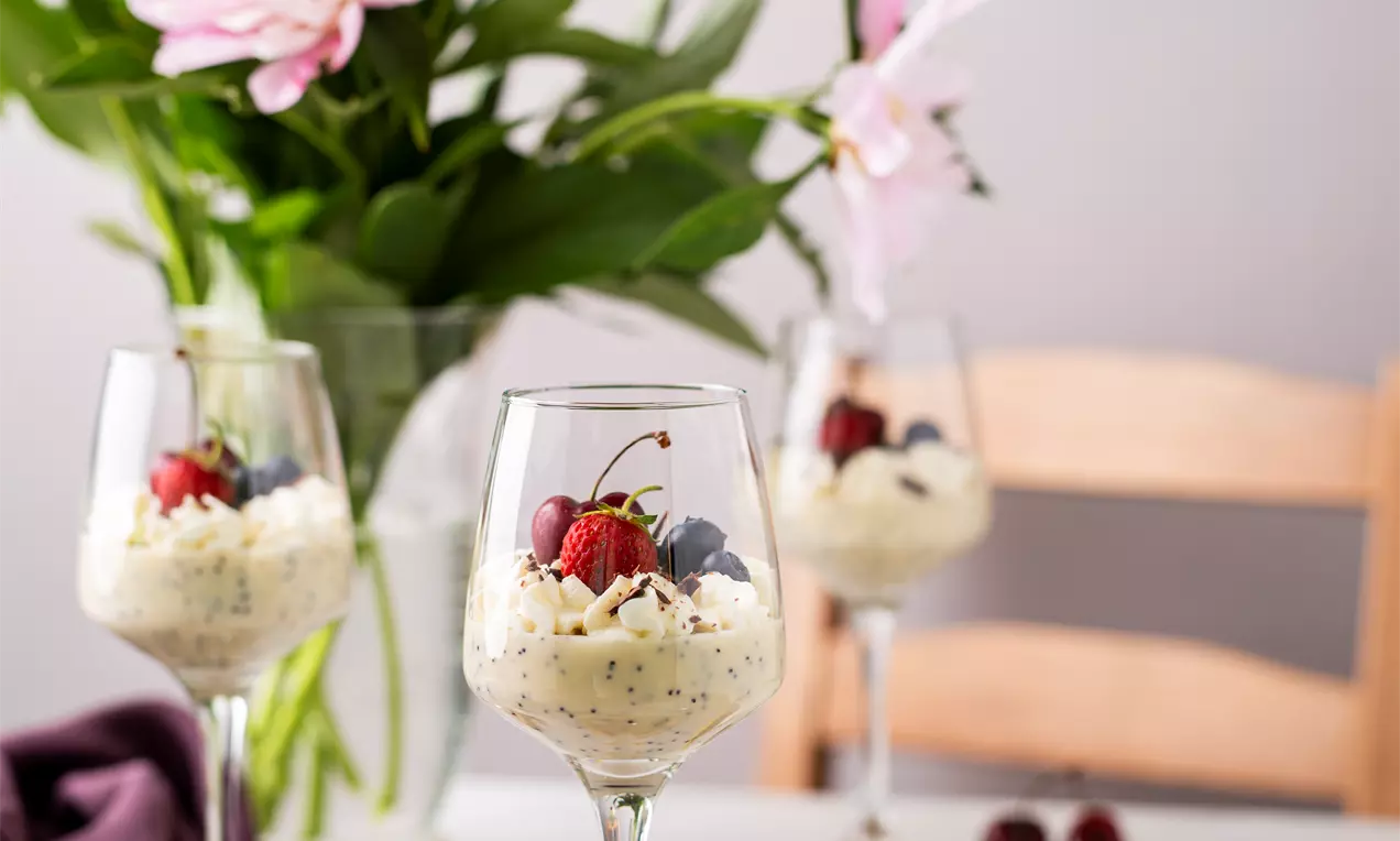 Eleganti deserti stikla vīna glāzēs ar gaišu krēmīgu pildījumu, melnām sēkliņām, rotāti ar ķiršiem un mellenēm, uz galda ar grāmatu un ziediem