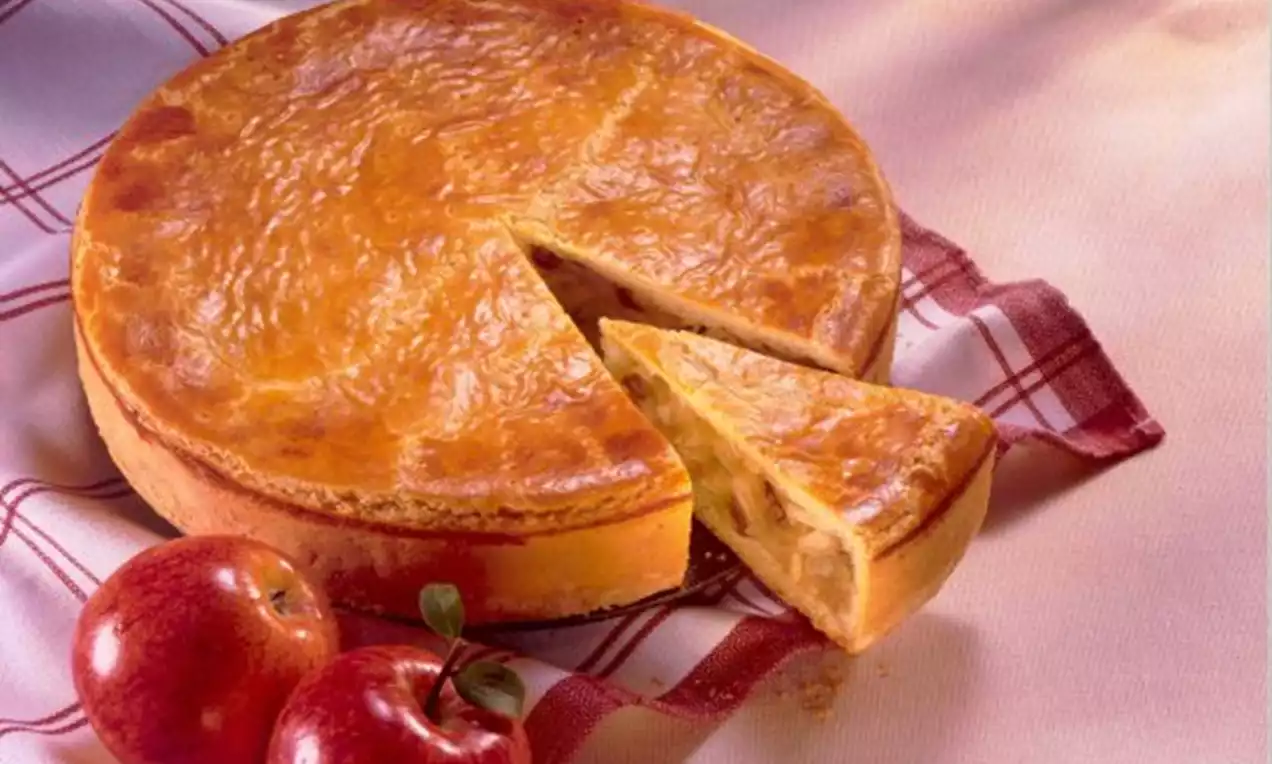 Goudbruine appeltaart met glanzende korst, een uitgesneden punt en zichtbare appelvulling, staand op een rood geruit doek naast rode appels