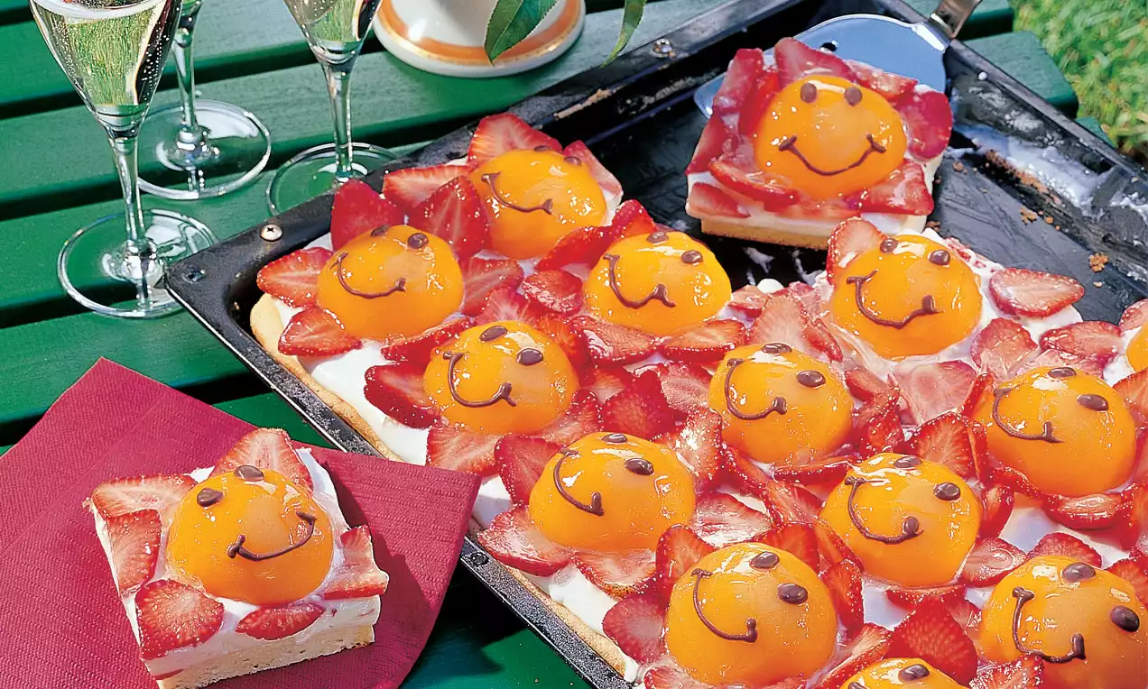 Rechteckiger Kuchen mit aufgeschnittenen Erdbeeren und ganzen Pfirsichhälften mit Gesichtern auf heller Creme auf einem Blech