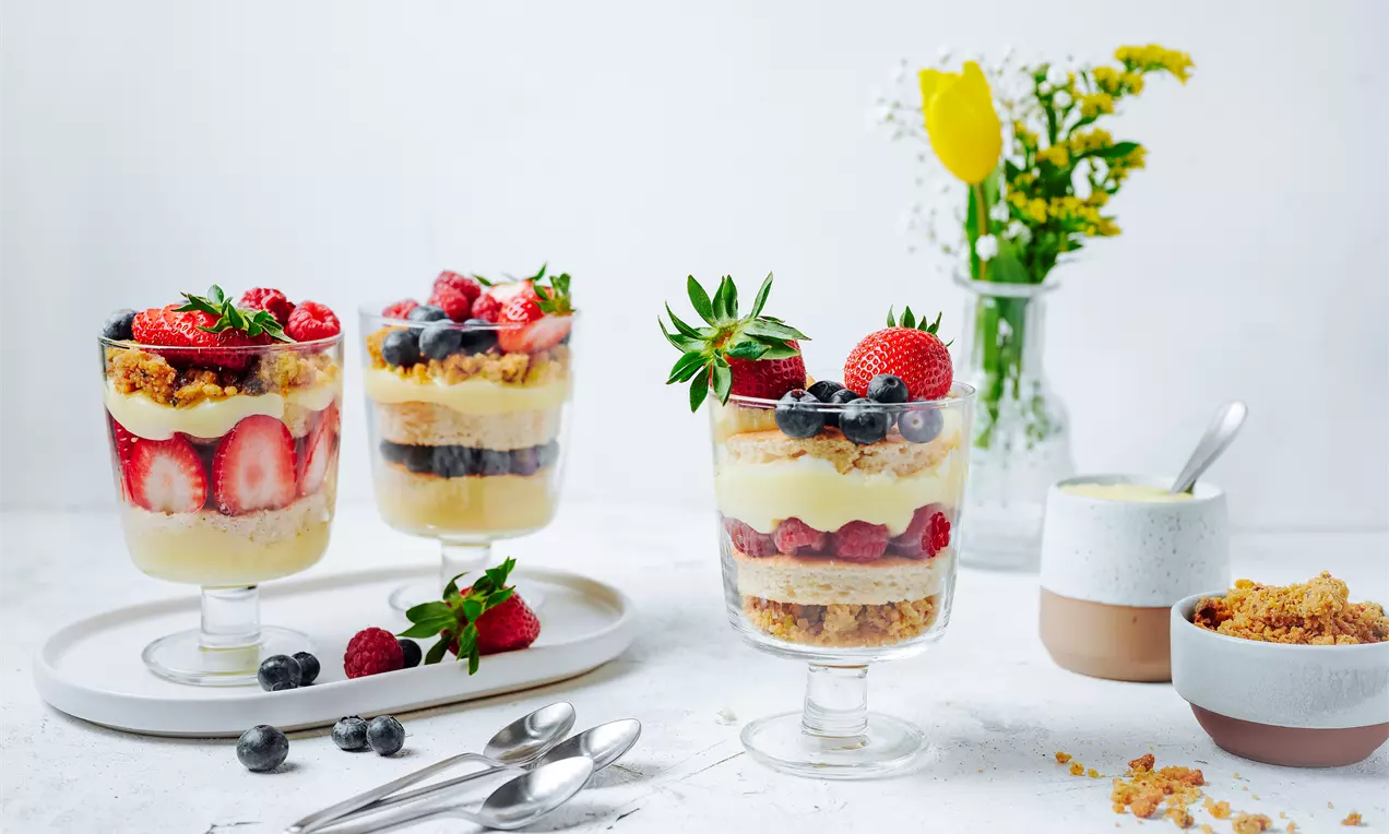 Пластове от велвего крем, златисти трохи и свежи плодове като ягоди и боровинки в стъклени чаши, сервирани с чинийки и лъжички