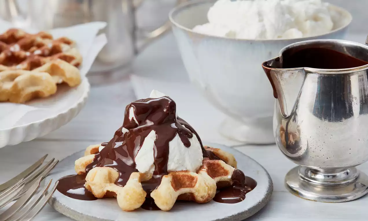 Goldbraune Lütticher Waffel auf grauem Teller, mit weißer Sahnehaube und glänzendem Schokoladenguss, umgeben von Sahneschüssel und Schokoladenkanne