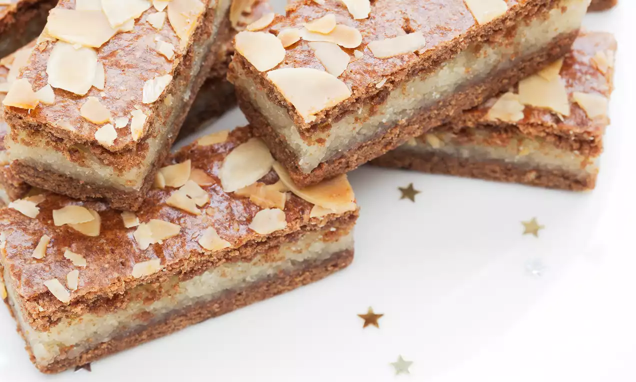 Rechthoekige plakken gevulde speculaas met een goudbruine korst, amandelschaafsel en een zachte laag op een wit bord met sterdecoratie