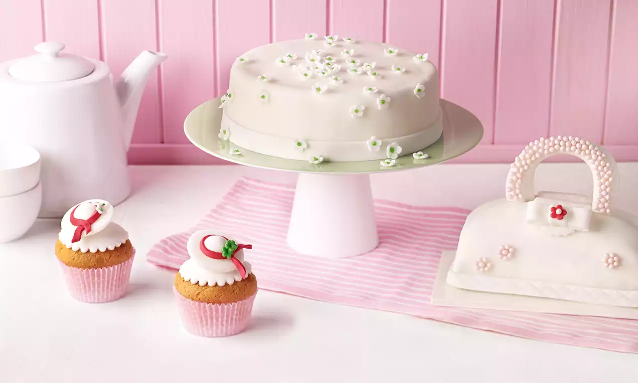 Eine runde Fondanttorte mit zarten weißen Blüten auf grüner Tortenplatte, daneben Cupcakes und eine Fondanttasche auf rosa Streifen-Unterlage