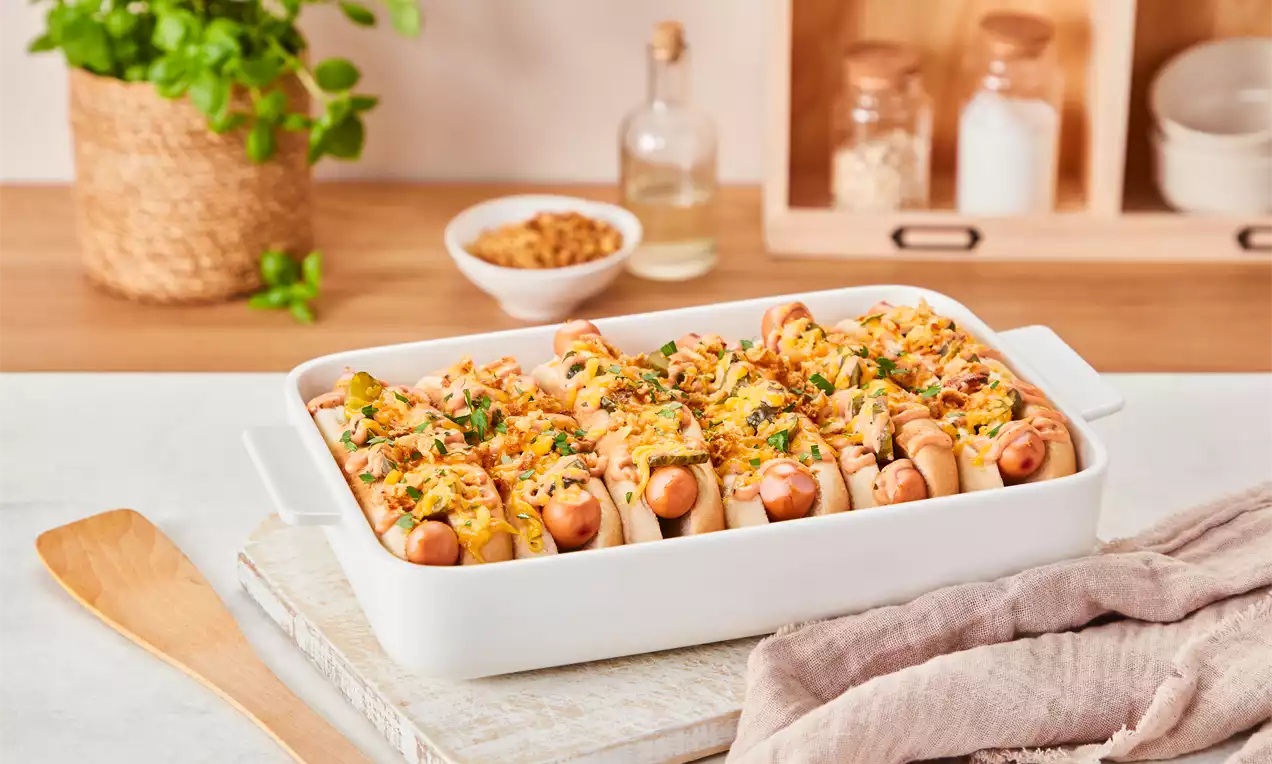 Goldbraune Hot Dogs in heller Auflaufform, mit geschmolzenem Käse, Petersilie und knusprigen Toppings, auf heller Küchenarbeitsfläche