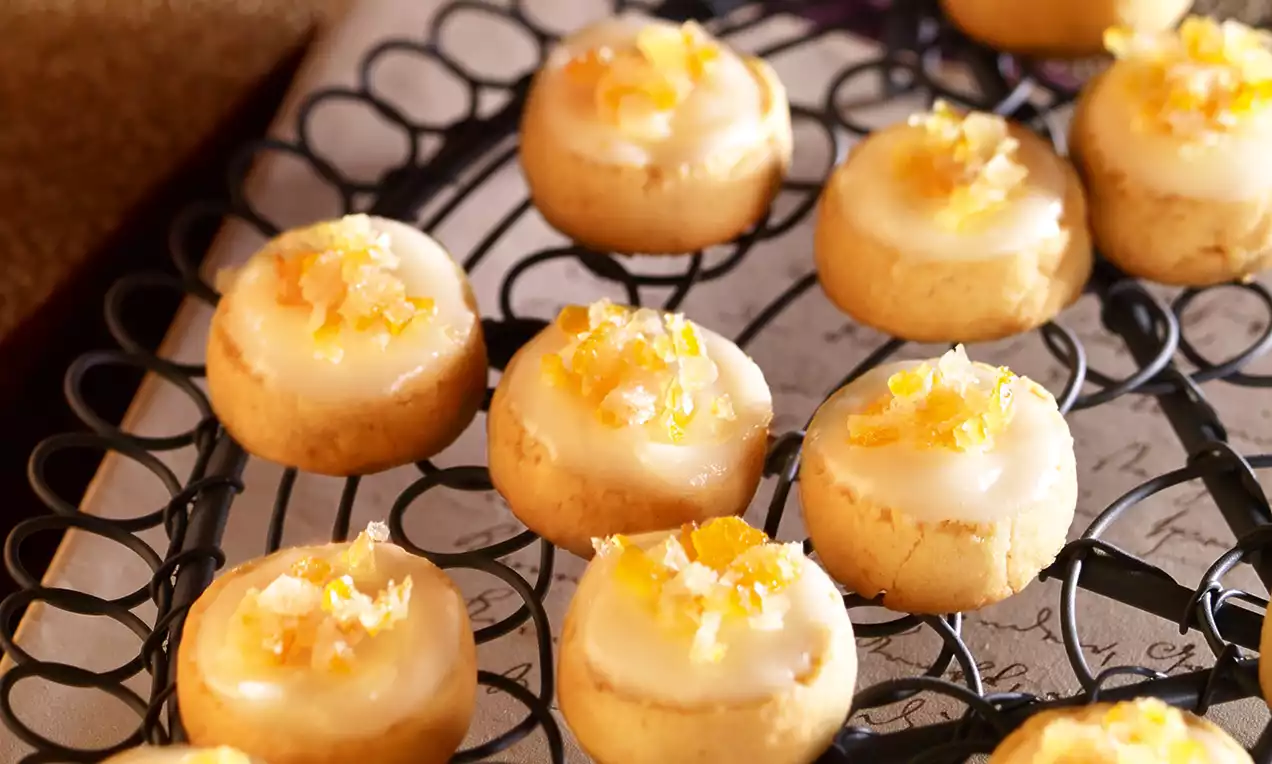 Kleine goldbraune Plätzchen mit glänzendem weißen Zuckerguss und fein gehackten orangefarbenen kandierten Früchten auf schwarzem Drahtgitter