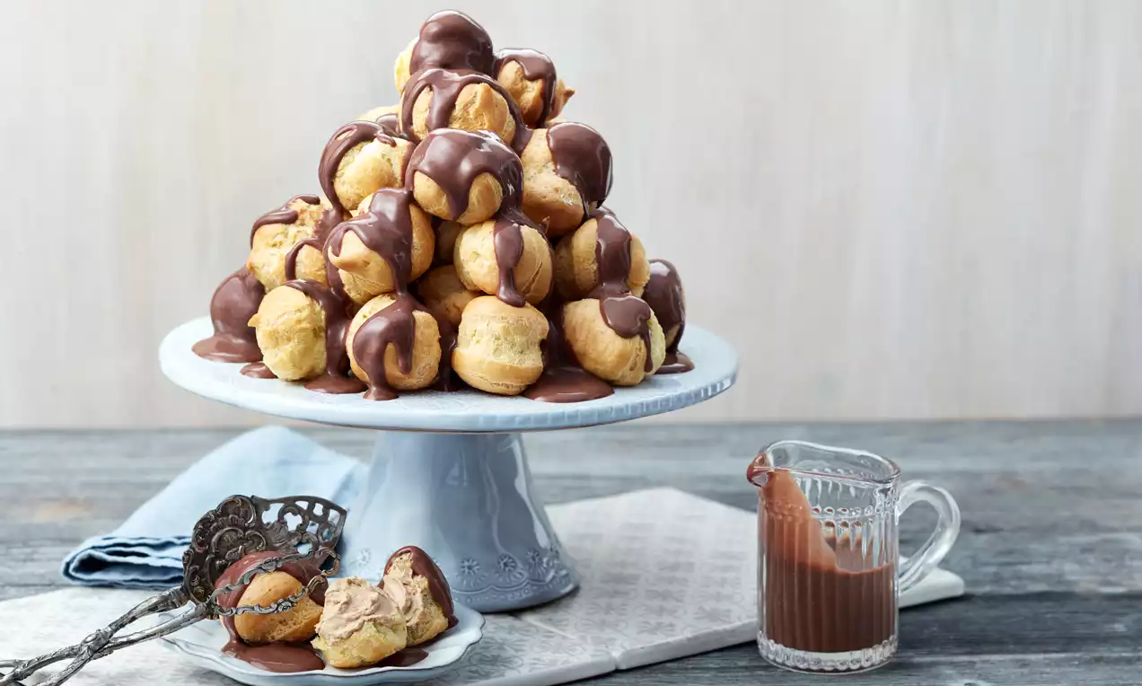 Profiteroler i pyramidform med gyllene, fluffig yta, fyllda med mockakräm, överdragna med chokladsås på vit tårtbricka bredvid en kanna chokladsås