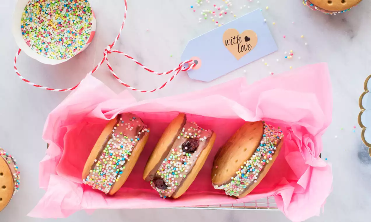 Keks-Sandwiches mit Schoko-Cranberry-Eis, bunte Streusel an den Rändern, auf rosa Papier in einer Box mit Geschenkanhänger und Dekoration