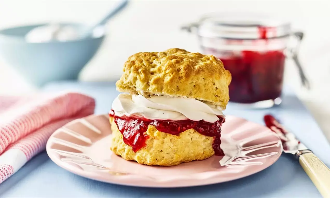 Златисто-кафяво скони с леко неравна повърхност, разделено и обилно запълнено с бял крем и яркочервено сладко върху розова чиния