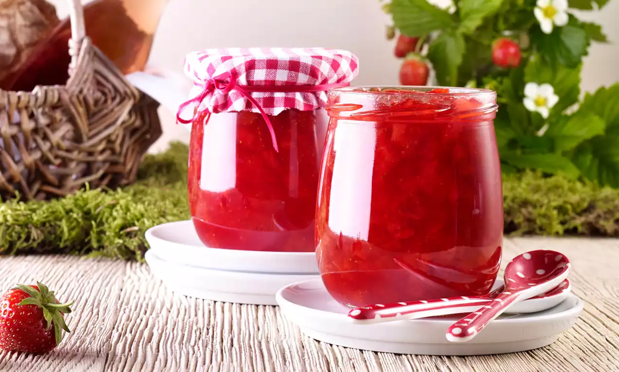 Erdbeerkonfitüre in gläsernen, rot glänzenden Gläsern mit karierten Deckeln, auf weißen Tellern mit rotem Löffel und frischen Erdbeeren