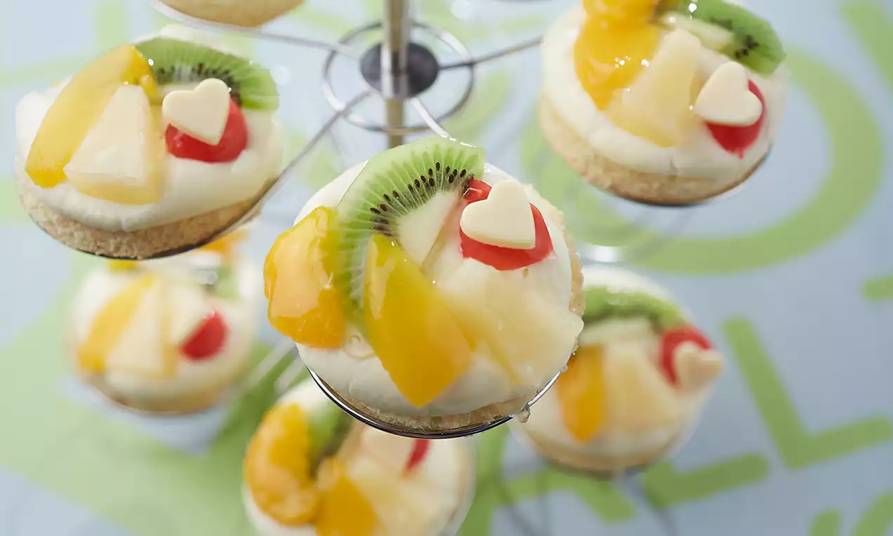 Runde Fruchttörtchen mit goldbraunem Boden, cremiger weißer Vanillecrème, bunten Früchten und Herzdekor auf einem silbernen Ständer