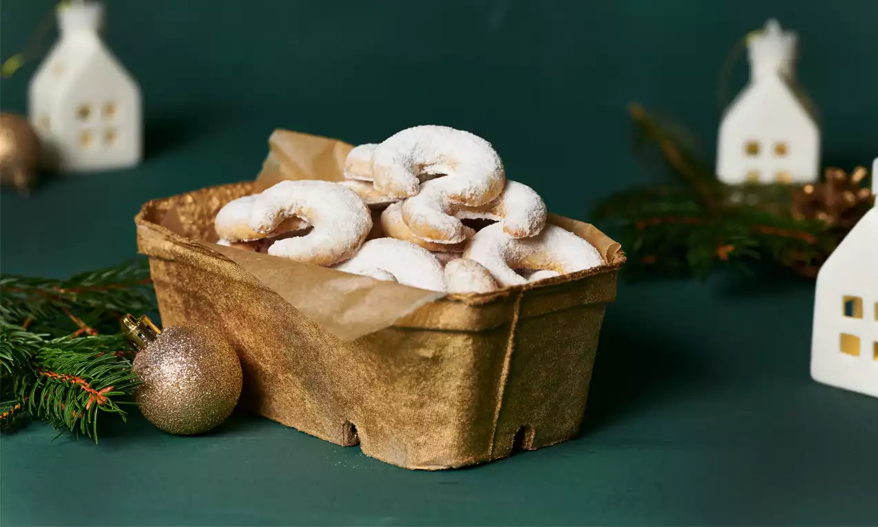 Goldbraune Vanillekipferl in geschwungener Hörnchenform, mit weißem Puderzucker bestäubt, auf braunem Papier in einer Kartonschale