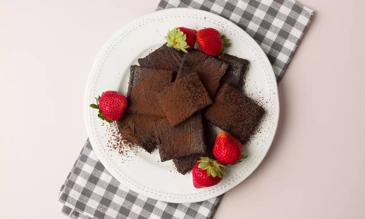 Brownies rectangulares de textura compacta y cobertura de cacao en polvo dispuestos en un plato blanco con fresas frescas