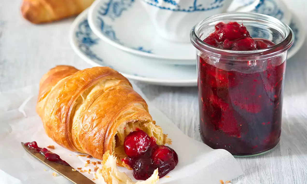 Zlatavý máslový croissant plněný lesklou červenou marmeládou vedle sklenice s džemem na bílém papíře, porcelánový šálek v modrém dekoru v pozadí